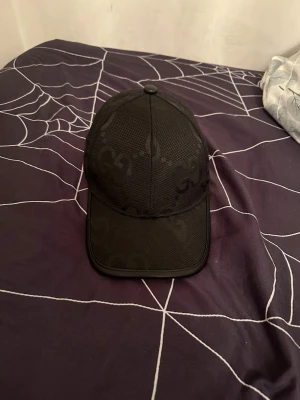 Svart keps från Gucci med GG-mönster - Snygg svart keps från Gucci med diskret GG-monogram över hela kepsen. Klassisk böjd skärm och justerbar passform. Tillverkad i Italien och har en exklusiv känsla med märkets ikoniska detaljer. Perfekt accessoar för att lyfta din streetwear-look.