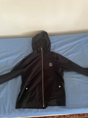 Svart vindjacka från Trapstar - Snygg svart vindjacka från Trapstar med huva och dragkedja framtill. Jackan har två fickor med tryckknappar och Trapstar-logga på bröstet samt stor text på ryggen. Perfekt för streetwear-stilen och enkel att matcha med olika outfits.