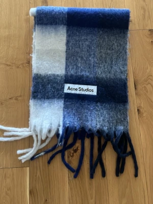 Acne Studios halsduk - Mjuk och fluffig halsduk från Acne Studios i rutigt mönster med blå och vita nyanser. Halsduken har långa fransar i ändarna och en tydlig logotyp påsydd. Perfekt accessoar för att addera färg och stil till din outfit för perfekt pris