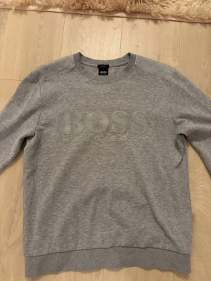 Grå sweatshirt från Hugo Boss - Snygg grå sweatshirt från Hugo Boss med stort BOSS-tryck på bröstet. Tröjan har rund halsringning, ribbade muddar och är tillverkad i mjukt bomullsmaterial. Perfekt för en avslappnad och stilren look.