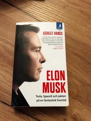 Elon Musk: Tesla, SpaceX och jakten - En pocketbok med vit framsida och stora röda bokstäver. Boken är skriven av Ashlee Vance och handlar om Elon Musk, Tesla och SpaceX. Omslaget har även ett citat och Månpocket-loggan i hörnet.