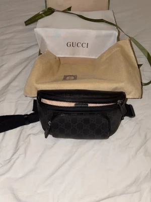 Gucci midjeväska - Snygg svart midjeväska med klassiskt GG-mönster. Väskan har justerbart band, dragkedja upptill och en extra ficka framtill. Tillverkad i skinn med detaljer i svart och silver. Perfekt för att bära det viktigaste med stil.