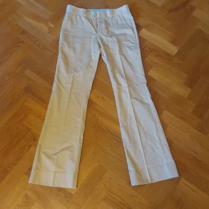 Beige kostymbyxor från J.Lindeberg - Säljer ett par stiliga beige kostymbyxor från J.Lindeberg. De har en klassisk rak passform med hög midja och är tillverkade i ett bekvämt bomullsmaterial. Byxorna har två bakfickor med knappar och stängs med en dragkedja och hyska. Perfekta för både kontoret och finare tillfällen!