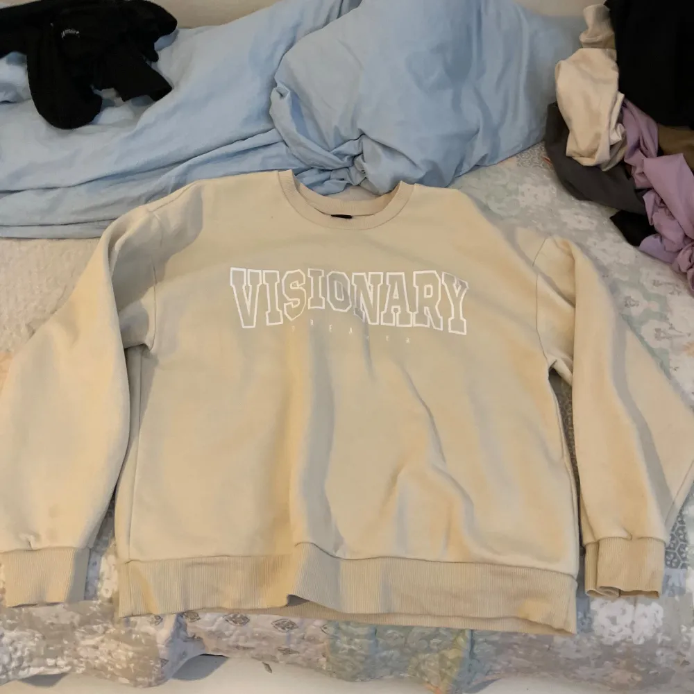 Säljer en snygg beige sweatshirt med texten 'VISIONARY' tryckt i vitt på framsidan. Tröjan har en klassisk rund hals och långa ärmar med ribbade muddar vid ärmslut och nederkant. Perfekt för en avslappnad stil!. Tröjor & Koftor.