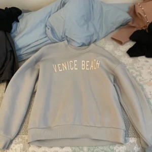 Grå sweatshirt med tryck 'Venice Beach' - Säljer en grå sweatshirt med texten 'Venice Beach' tryckt på framsidan. Tröjan är supermjuk och perfekt för en avslappnad stil. Den har långa ärmar och ribbade muddar vid ärmslut och nederkant. Perfekt för höst och vår!