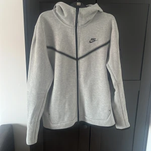Grå Nike Tech fleece hoodie  - Säljer en grå Nike Tech hoodie i storlek M. Köpt för ungefär 4-5år sen när jag var yngre (runt 15-16) och har aldrig använt den. Köpt från JD sports men föräldrarna hittar ej kvittot. (Original pris 1250kr)