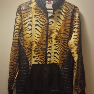 Tuff tigerjacka med dragkedja - Säljer en riktigt cool jacka med tigerprint. Den har en dragkedja framtill och quiltade svarta paneler på sidorna och ärmarna. Perfekt för att sticka ut i mängden! 🐯