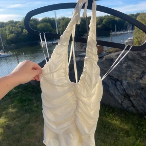 Vit klänning med rynkdetaljer - Säljer en superfin vit topp med rynkdetaljer och med knytning baktill. Toppen har en tight passform och är gjord i ett mjukt och bekvämt material mot kroppen, då det är en body knäpper man den med spännen