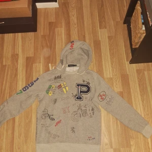Grå hoodie med tryck från polo ralph lauren - Priset är inte satt i sten kom gärna med förslag är villig att sänka!                           Säljer en grå hoodie från Ralph Lauren med olika tryck och en stor bokstav 'P' på framsidan . Hoodien har en justerbar huva och långa ärmar. Perfekt för en avslappnad stil. (Skick 9-10)