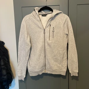 Grå huvtröja från Sail racing - Säljer en stilren grå huvtröja från Sail racing. Tröjan har en dragkedja framtill och en praktisk huva. Perfekt för en avslappnad stil.