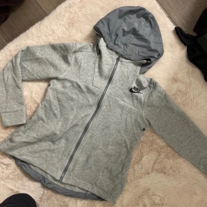 Grå huvtröja från Nike - Säljer en grå huvtröja från Nike med dragkedja och huva. Tröjan är tillverkad i en blandning av bomull och polyester, vilket gör den både bekväm och hållbar. Perfekt för en avslappnad stil. Finns en liten fläck på höger arm