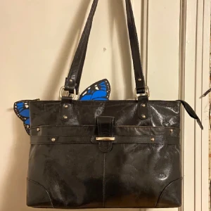 Svart axelväska med fjärilsdetalj - Snygg svart axelväska. Väskan har en praktisk dragkedja och ett stilrent spänne framtill. Perfekt för att ge din outfit en extra touch.