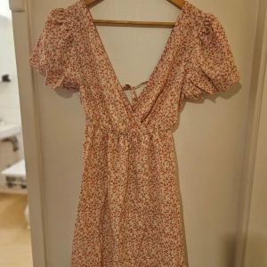 Blommig klänning från Romwe - Söt blommig klänning från Romwe i ljusa toner med korta puffärmar och v-ringning. Klänningen har ett knytband i ryggen och ett fint blommönster i rosa och grönt.