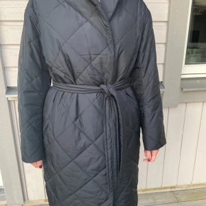Svart quiltad kappa från Arket - Snygg svart quiltad kappa från Arket med knytbälte i midjan. Perfekt för kyligare dagar med sin långa design och långa ärmar. En stilren och tidlös jacka för alla tillfällen.