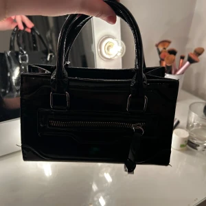Svart handväska med dragkedja - Snygg svart handväska i glansigt material med praktisk dragkedja framtill. Perfekt för att ge en stilren touch till din outfit. Enkel att bära med sina dubbla handtag.