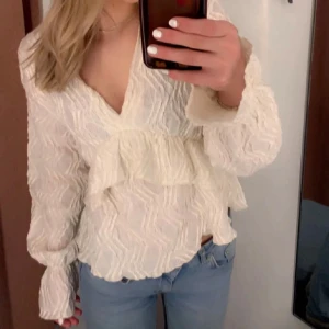 Vit blus med volangdetaljer - Säljer en söt vit blus med volangdetaljer och v-ringning. Blusen har långa ärmar och ett lätt strukturerat mönster. Perfekt för en stilren look.