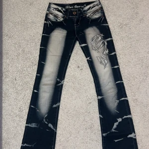 Mörkblå jeans med drakbroderi - Snygga mörkblå jeans från Blue One med unikt drakbroderi på benet och bakfickorna. Jeansen har en cool tvättad look med ljusa partier och slitningar. De har dragkedja och knapp framtill samt dekorativa dragkedjor vid fickorna.
