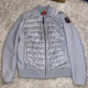 Grå jacka från Parajumpers - Snygg grå jacka från Parajumpers med quiltade detaljer och dragkedja framtill. Jackan har en patch på ärmen och ribbade muddar. Perfekt för kyligare dagar. Den är väldigt bra skick. Har aldrig använts. Jackan kan passa S. Den är äkta. Priset kan diskuteras!