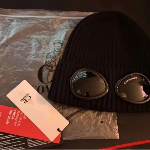 Svart ribbad mössa med glasögon från C.P. Company - Snygg svart ribbad mössa från C.P. Company med inbyggda glasögon. Perfekt för en unik och stilren look. Mössan är både praktisk och trendig.