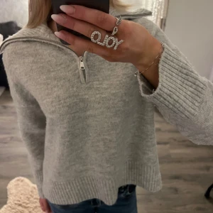 Grå stickad tröja med dragkedja - Mysig grå stickad tröja med dragkedja vid halsen. Perfekt för kyliga dagar med sin ribbade struktur och långa ärmar. En stilren och bekväm tröja som passar till det mesta.