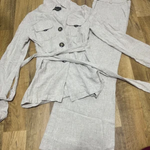 Beige linnekostym med bälte - Elegant beige kostym i linne med kavaj och matchande byxor. Kavajen har knappar och ett bälte i midjan för en snygg passform. Perfekt för vår och sommar. Priset går att diskuteras vid snabb affär