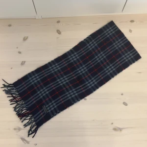 Halsduk från Burberry - Säljer en klassisk rutig halsduk från Burberry i mörkblått med inslag av grått och rött. Halsduken har fransar i ändarna och är perfekt för att hålla dig varm under kyliga dagar. Ett tidlöst och stilrent val för garderoben.