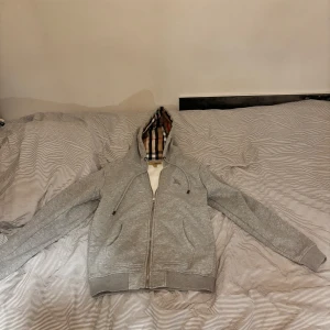 Grå hoodie från Burberry - Säljer en stilren grå hoodie från Burberry med klassiskt rutigt mönster i huvan. Tröjan har dragkedja och broderad logga på bröstet. Perfekt för en avslappnad och trendig look.(🐼)
