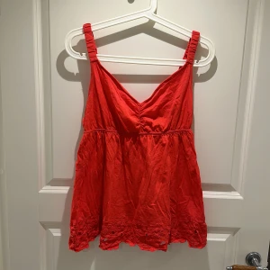 Rött linne - Säljer ett snyggt rött linne med smala axelband och v-ringning. Toppen har en luftig passform och fina broderade detaljer längst ner. Perfekt för varma sommardagar!