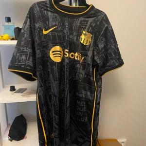 Svart FC Barcelona fotbollströja - Snygg svart FC Barcelona fotbollströja från Nike med gula detaljer och tryck. Tröjan har korta ärmar och ett unikt mönster som täcker hela tyget. Perfekt för fans av klubben!