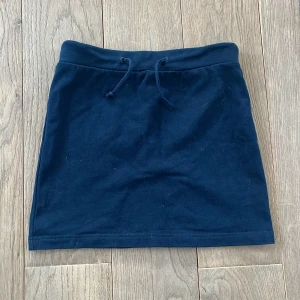 Mörkblå kjol med knytband - Snygg mörkblå kortkjol med knytband i midjan och en liten ficka bak. Perfekt för en avslappnad stil och svalt på sommaren. Kjolen är gjord av 100 % bomull.