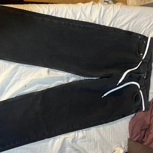 Svarta jeansbyxor från Junkyard (District Fortysix) - Snygga svarta jeansbyxor från District med rak passform. Byxorna har en klassisk design med fem fickor och en vit snörning i midjan för extra stil. Perfekta för en avslappnad look.