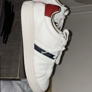Vita sneakers från Gant - Snygga vita sneakers från Gant med röda och blå detaljer. Skorna har en klassisk design med snörning och en röd sula. Perfekta för en stilren look. Passar in med dagens stil, kommer inte med original boxen. Ord pris 1500kr, använd 5-10 gånger 