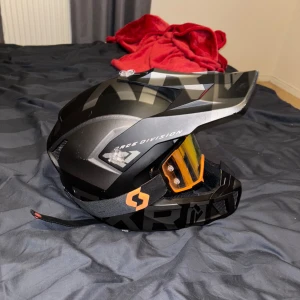 Svart hjälm med orange visir - Snygg svart hjälm med orange visir och Race Division. Hjälmen har en justerbar rem och bekväm vaddering inuti. Har små skavanker men inget man lägger märke till. 