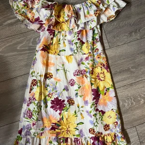 Blommig offshoulder klänning - Charmig offshoulder klänning med ett färgglatt blommönster i gult, lila och grönt på en vit bakgrund. Klänningen har volangdetaljer vid axlarna och en bra kropps passform, perfekt för sommarens varma dagar.🌸🪻 Storlek S men passar även XS🌾