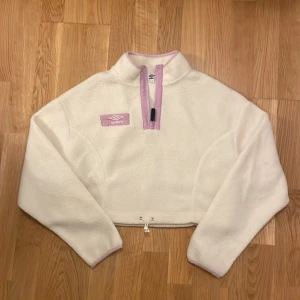 Vit fleece tröja från Umbro - Säljer en mysig vit fleece tröja från Umbro med ljuslila detaljer. Tröjan har en halv dragkedja framtill och en justerbar dragsko i nederkanten. Perfekt för kyliga dagar!