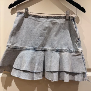 Ljusblå jeanskjol med volang - Trendig ljusblå jeanskjol från H&M med volangdetaljer och rå kant. Kjolen har en dragkedja på sidan för enkel på- och avtagning. Perfekt för en avslappnad stil.