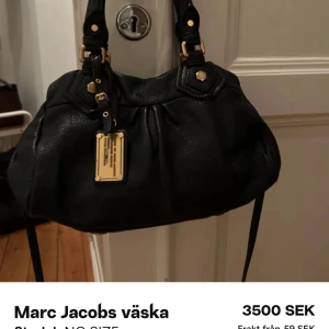 Svart väska Marc Jacobs - Snygg svart handväska från Marc Jacobs i skinn med gulddetaljer och en elegant design. Väskan har dubbla handtag och en dekorativ etikett framtill. Perfekt för att ge din outfit en lyxig touch.