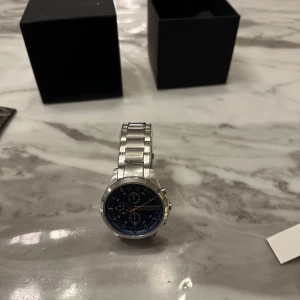 Silverfärgad klocka från Armani Exchange - Snygg klocka från Armani Exchange med en stilren silverfärgad länkarmband och en mörk urtavla. Urtavlan har flera små visare och orange detaljer som ger en sportig känsla. Perfekt för den som vill ha en kombination av stil och funktion.