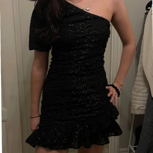 Svart glittrig klänning - Snygg svart glittrig klänning med oneshoulder-design och kort längd. Klänningen har en volangdetalj vid fållen och är perfekt för fest!!🥂🥂