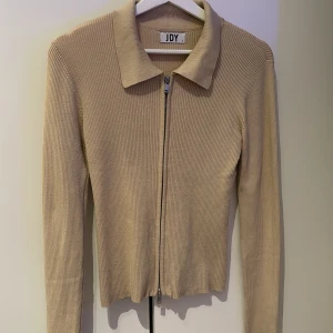 Beige ribbad kofta från JDY - Snygg beige kofta från JDY med ribbad struktur och dragkedja framtill. Den har en klassisk krage och långa ärmar, perfekt för lager på lager-stil. En stilren och mångsidig tröja för alla tillfällen. Kedjan går att justera både uppifrån och nedifrån.
