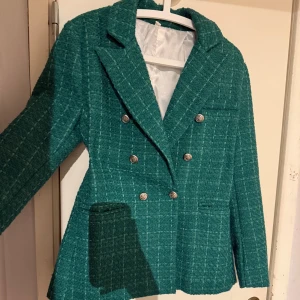 Grön strukturvävd kavaj med gulddetaljer - Snygg grön kavaj som är figursydd och har dubbla rader av dekorativa knappar. Perfekt för en stilren look. Kavajen har en klassisk krage och långa ärmar. Endast använd vid ett fåtal tillfällen