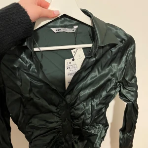 Grön satinblus från Zara - Snygg grön satinblus från Zara med knappar framtill. Blusen har en elegant glans och en figurnära passform som ger en stilren look. Perfekt för att lyfta vilken outfit som helst. Prislappen är på, storleken är i xs! Pris kan diskuteras 