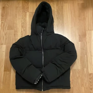 Svart pufferjacka från Le Temps des Cerises - Säljer en svart pufferjacka från Le Temps des Cerises. Jackan har en oversized passform och är utrustad med en dragkedja och huva. Perfekt för att hålla dig varm och stilren under kyliga dagar.