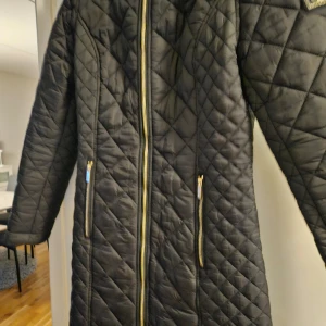 Svart quiltad kappa från Cavalleria Toscana - Snygg svart quiltad kappa från Cavalleria Toscana med dragkedja och huva. Perfekt för kyliga dagar med sin långa design och stilrena look. Kappan har en elegant passform och är idealisk för både vardag och speciella tillfällen.