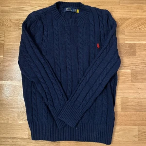 Polo Ralph Lauren - Säljer en stilren mörkblå kabelstickad tröja från Polo Ralph Lauren med det klassiska röda logotypbroderiet på bröstet. Tröjan har långa ärmar och ribbade muddar vid ärmslut och nederkant. Perfekt för kyligare dagar!