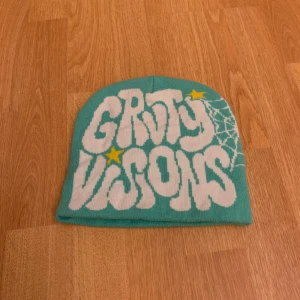 Turkos mössa med tryck - Cool turkos mössa med texten 'GRAVITY VISIONS' i vitt och en stjärna i gult. Har ett spindelnätsmönster på sidan. Perfekt för att sticka ut i mängden!