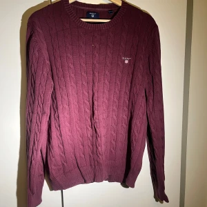 Vinröd kabelstickad tröja från GANT - Säljer en stilren vinröd kabelstickad tröja från GANT. Tröjan har en rund halsringning och långa ärmar, perfekt för kyligare dagar. Den är prydd med GANT-loggan på bröstet. Passar både till vardags och mer uppklädda tillfällen.