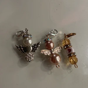Änglaformade berlocker med pärlor 10 kr styck  - Tre änglaformade berlocker med pärlor i olika färger. En har silverfärgade vingar och vita pärlor, en har guldfärgade vingar och rosa pärlor, och en har guldfärgade detaljer med gula pärlor och ett hjärtmönster. Perfekta som personliga smycken eller dekorationer.