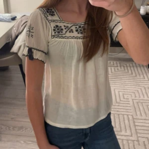 Vit blus med broderi - Fin vit blus med svart broderi på bröstet och ärmarna. Blusen har korta ärmar med små volanger och en lös passform. Tröjan har en liten defekt, den har tappat lite svart färg på baksidan av tröjan. Annars mycket bra skick och består av 100% bomull. 