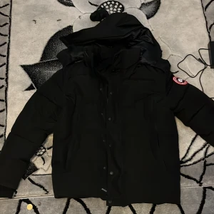 Svart dunjacka från Canada Goose - Säljer en svart dunjacka från Canada Goose med deras ikoniska märke på ärmen. Jackan har en hög krage och långa ärmar, perfekt för kalla vinterdagar. Den är både stilren och funktionell med dragkedja och knappar framtill.